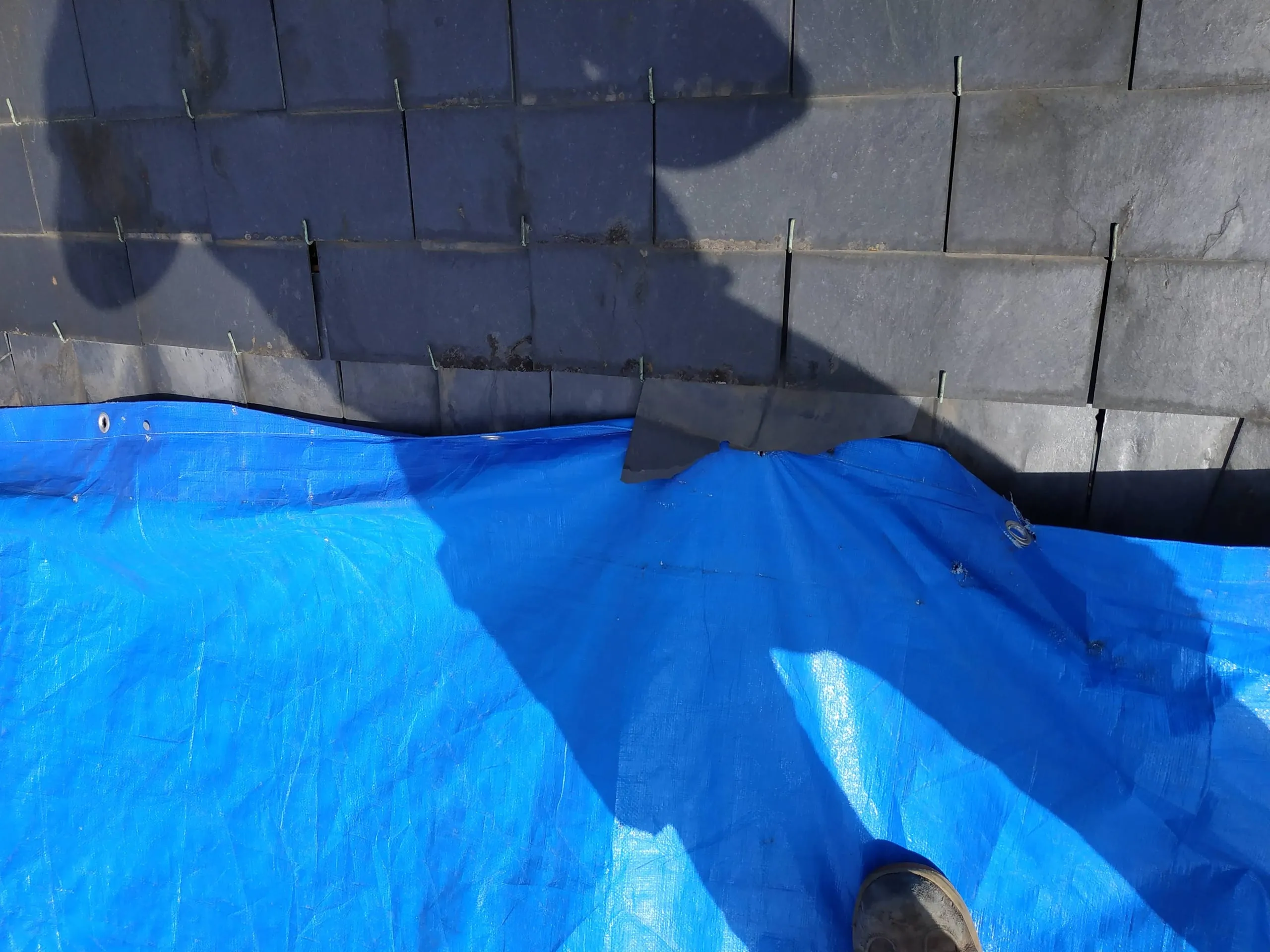 Toiture en ardoise partiellement protégée par une bâche bleue près d’une cheminée