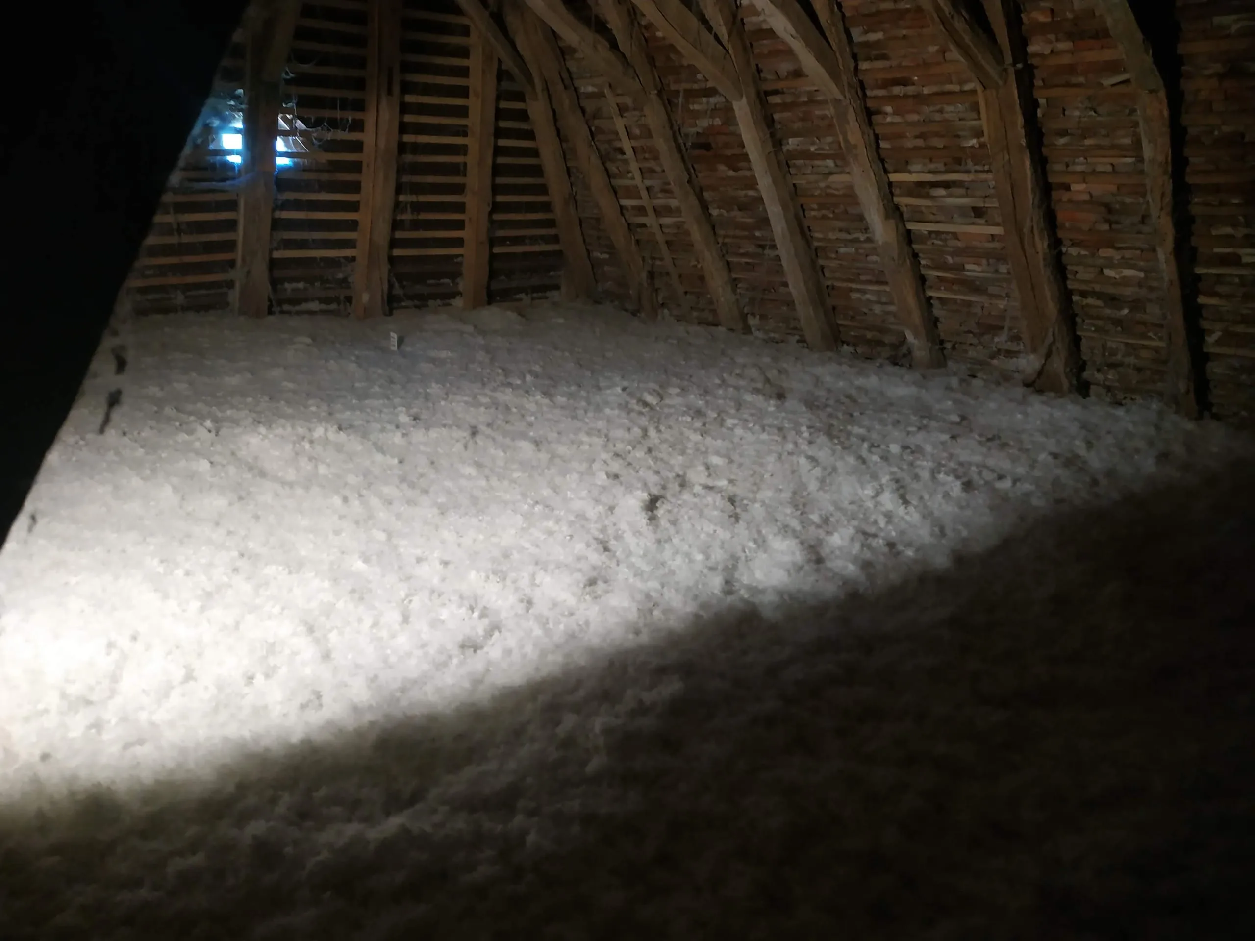 Isolant soufflé blanc dans des combles sous une charpente ancienne en bois et briques