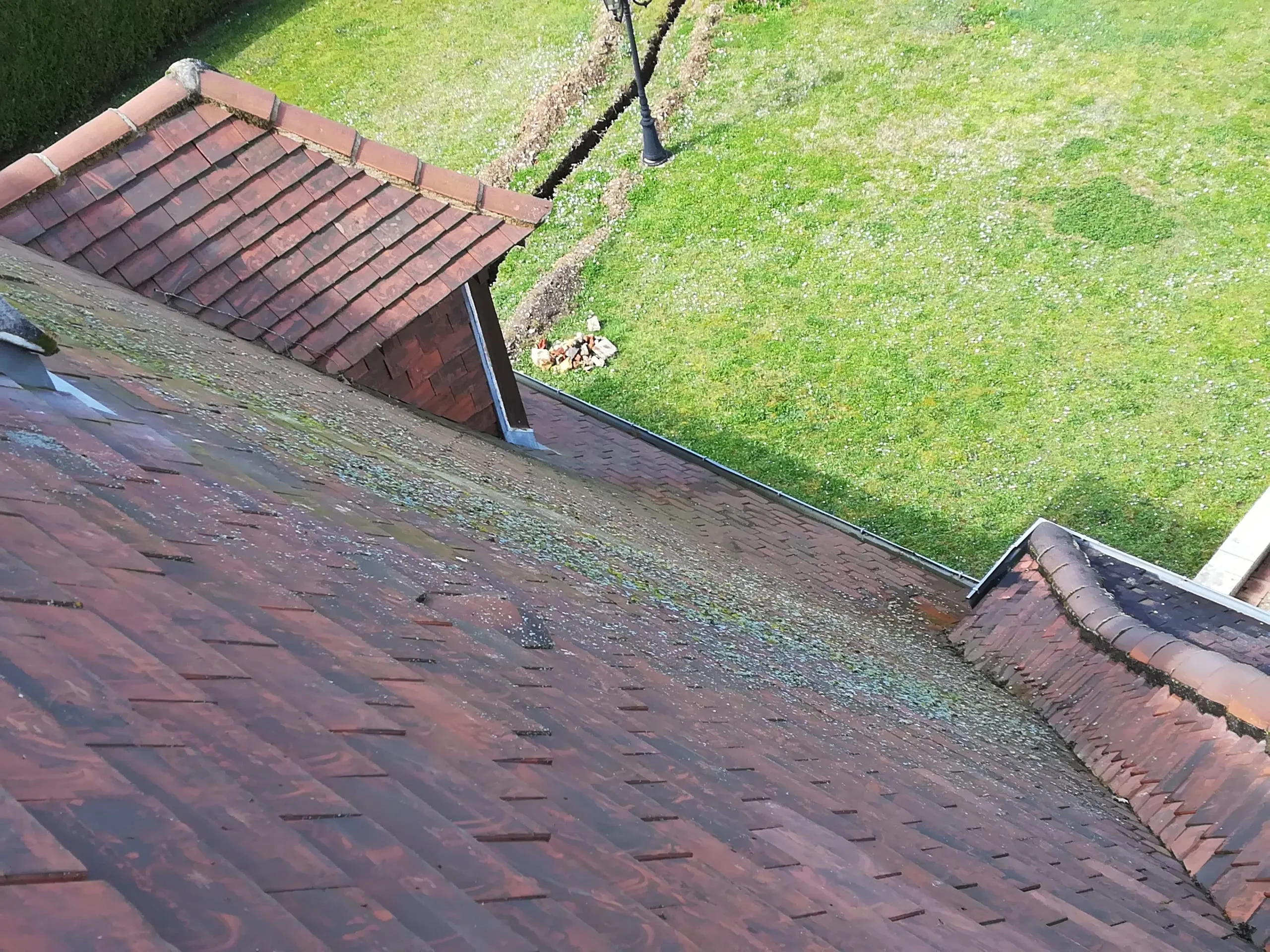 Vue en plongée de plusieurs pans de tuiles anciennes marqués par la mousse, avec des jonctions et une pelouse